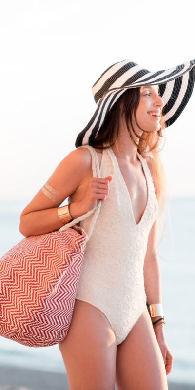 Young woman in hat and swimsuit walking with beach bag near the ocean during the summer vacation